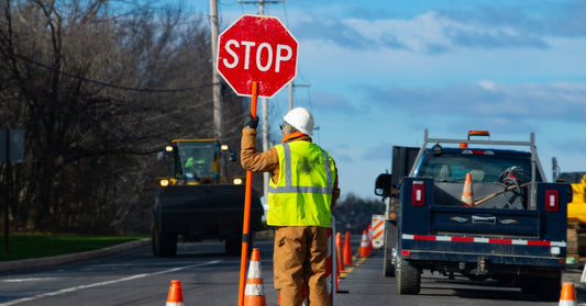 A road crew member stands in the street in a hi-vis mesh safety vest as they hold a stop sign to direct traffic.