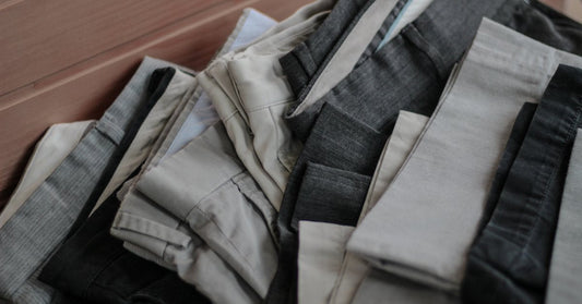 A pile of FR rain pants folded up on the floor. The pants are a mix of gray, denim, and dark khaki.