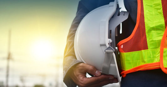 A close-up of a construction worker wearing a safety vest as they hold their white hard hat with their wrist.