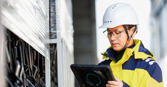A data center professional wears a cold-weather coat, a white hard hat, and holds a tablet as they work on a panel.
