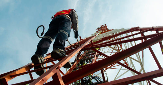 A tower work professional wearing a safety harness, hard hat, and fall protection device stands on the side of a tower.