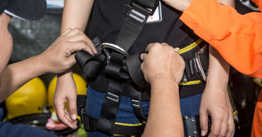 One professional helps another professional get into fall protection gear as they tighten the hoist and buckles.
