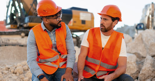 Two construction workers sit on the side of the job in hi-vis orange safety vests and matching orange hard hats.