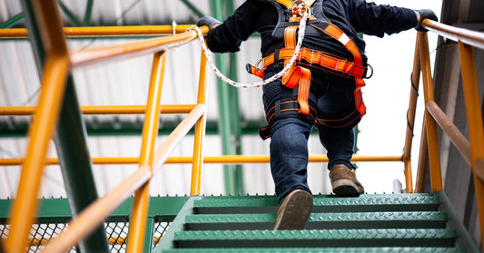 An industrial employee climbs a set of green stairs in an orange safety harness. There's a cord attached to the railing.