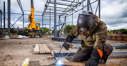 A welder working outside in full FR gear. They have a helmet on as they take the welder to pieces of metal.