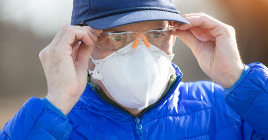 A close-up of a man in a blue jacket and hat outside at a jobsite as he adjusts his safety glasses and wears a mask.