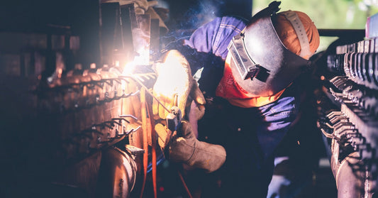 A professional welder working closely with a welding gun as sparks fly toward them. They wear a face shield and gloves.
